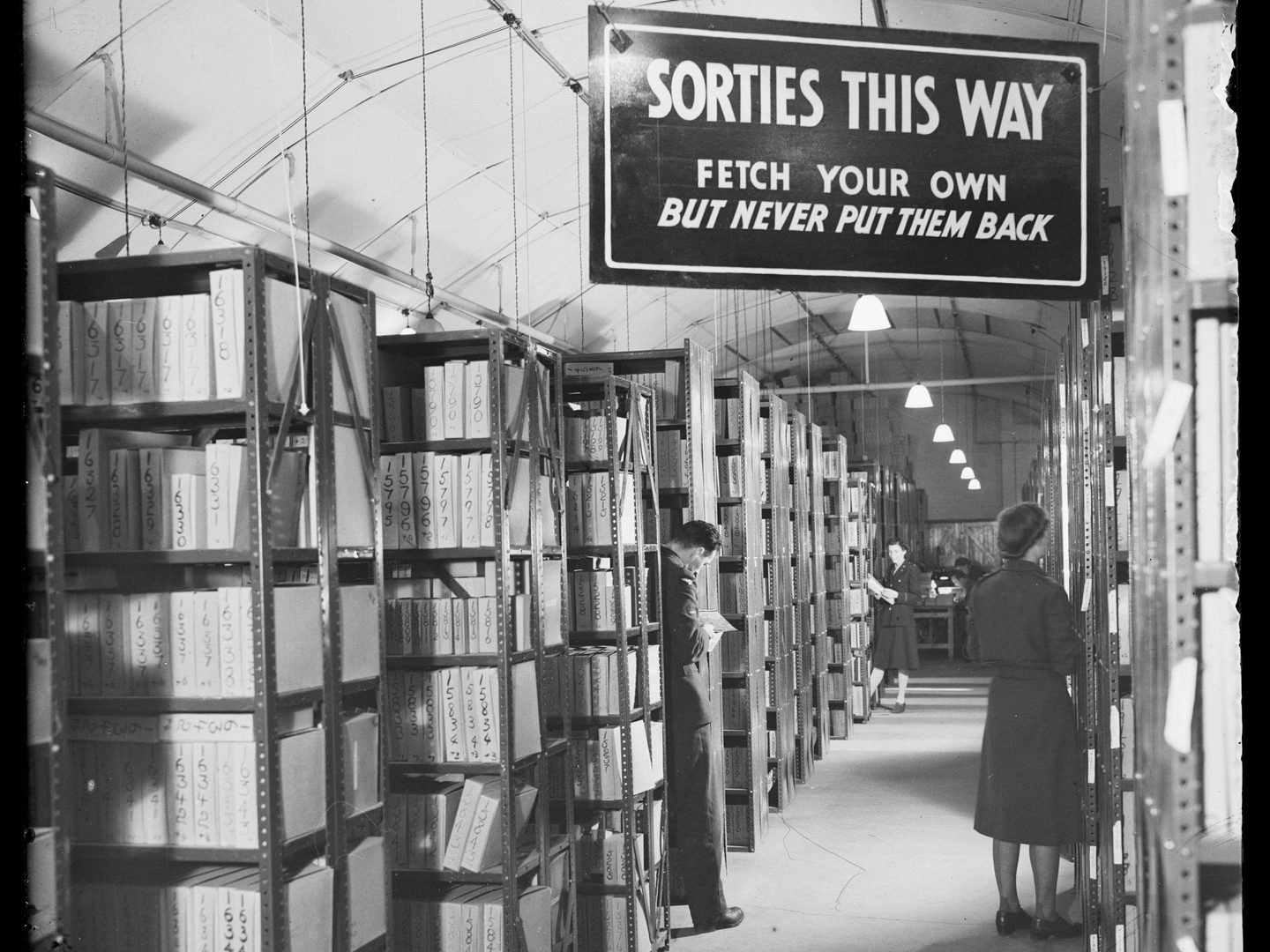 A black and white photo of a room filled with metal racking. Every rack is filled with numbered cardboard boxes. A man and woman in uniform stand with their backs to the camera. A sign reads ‘Sorties This Way: fetch your own but never put them back’.