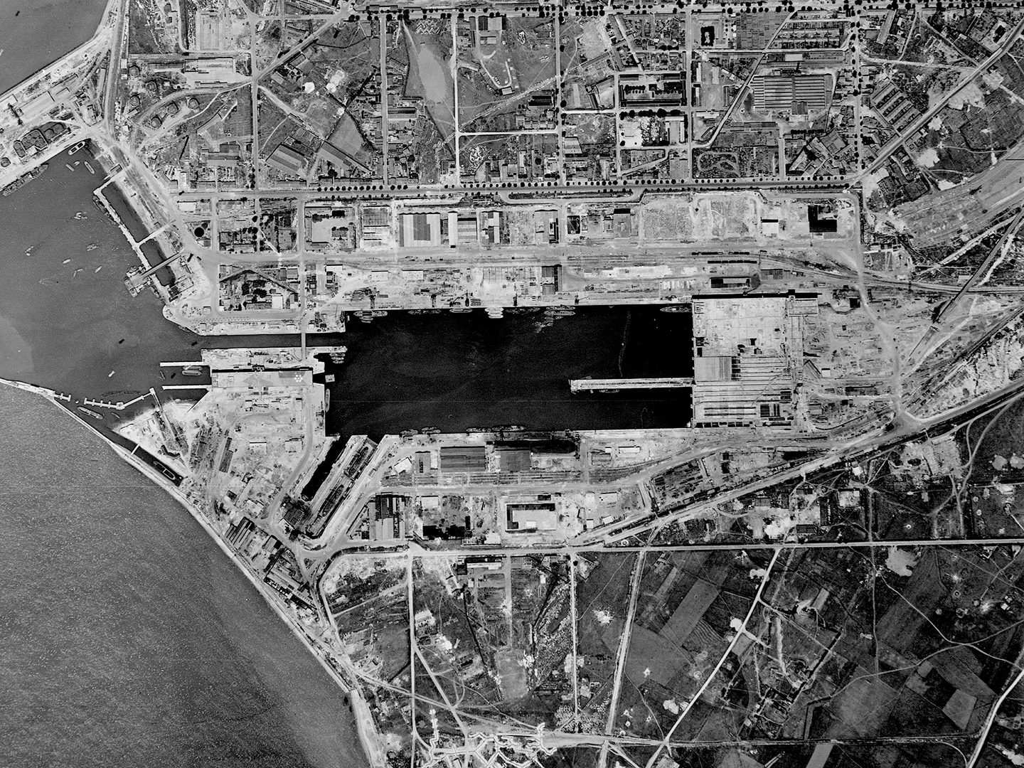 A historic black and white vertical aerial photo of a city with a rectangular area of dark water in the middle of it. To the left of the image, the city ends at a coastline.