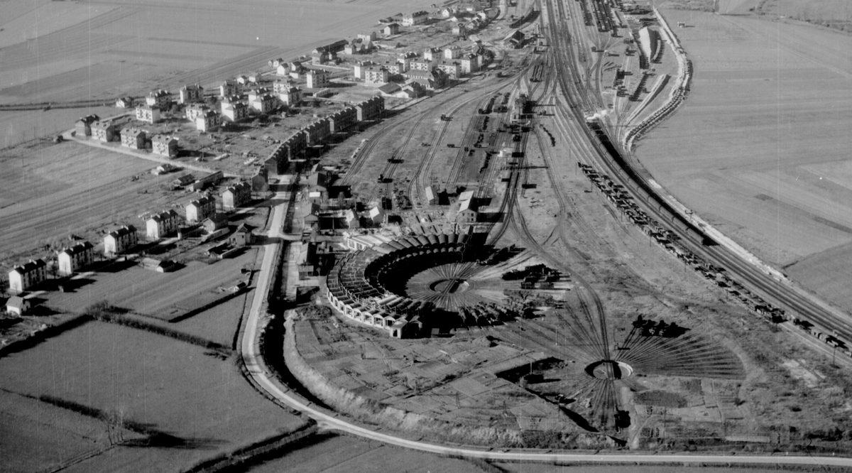 A black and white oblique aerial photo of a circular railway engine shed. A railway line stretches across the right-hand side of the image and the houses of a village are visible to the left.