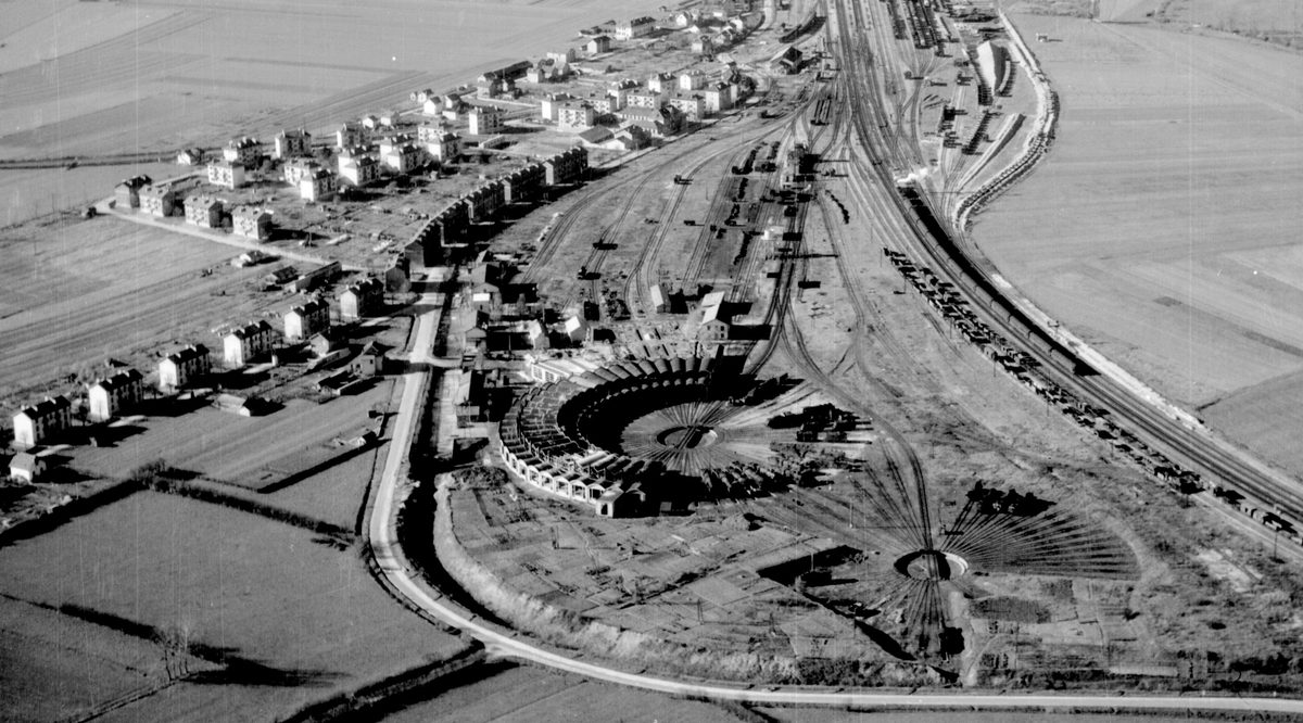 A black and white oblique aerial photo of a circular railway engine shed. A railway line stretches across the right-hand side of the image and the houses of a village are visible to the left.