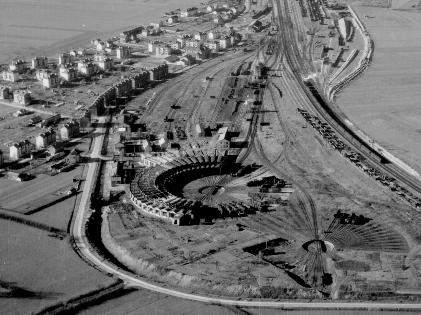 A black and white oblique aerial photo of a circular railway engine shed. A railway line stretches across the right-hand side of the image and the houses of a village are visible to the left.
