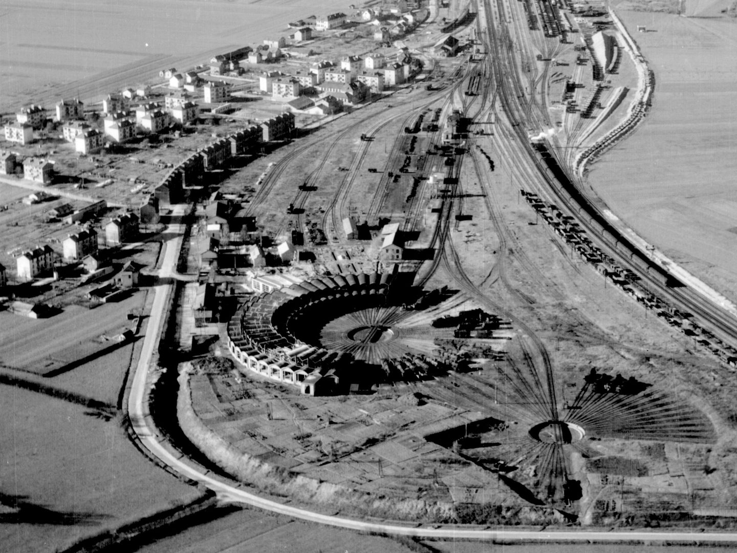 A black and white oblique aerial photo of a circular railway engine shed. A railway line stretches across the right-hand side of the image and the houses of a village are visible to the left.