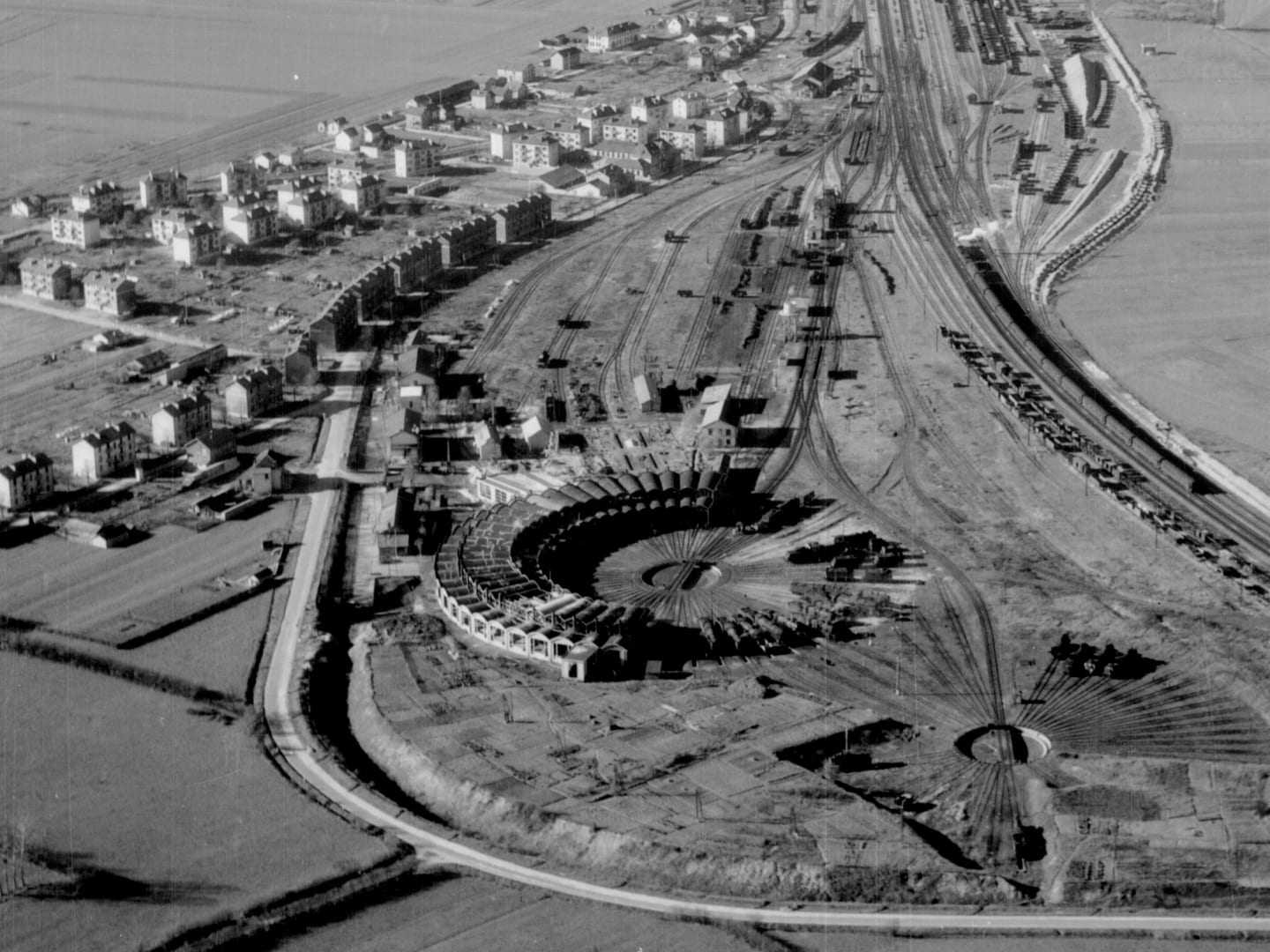 A black and white oblique aerial photo of a circular railway engine shed. A railway line stretches across the right-hand side of the image and the houses of a village are visible to the left.