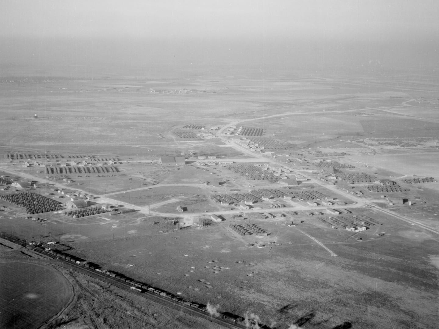 A black and white oblique aerial photo of a large encampment surrounded by fields. A railway line runs across the bottom edge of the image with a train passing along it.