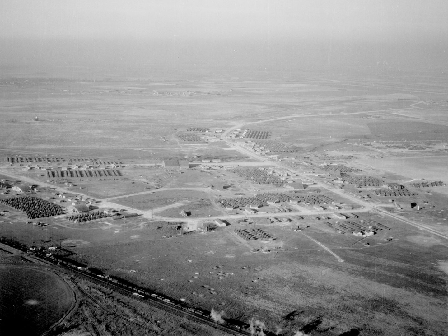 A black and white oblique aerial photo of a large encampment surrounded by fields. A railway line runs across the bottom edge of the image with a train passing along it.