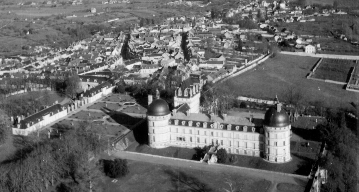 A historic black and white oblique aerial photo of a French chateau. It has white walls and 2 circular towers joined by a long wing. A second wing runs at rightangles from one of the towers to a square tower. It is surrounded by countryside with a town in the background.