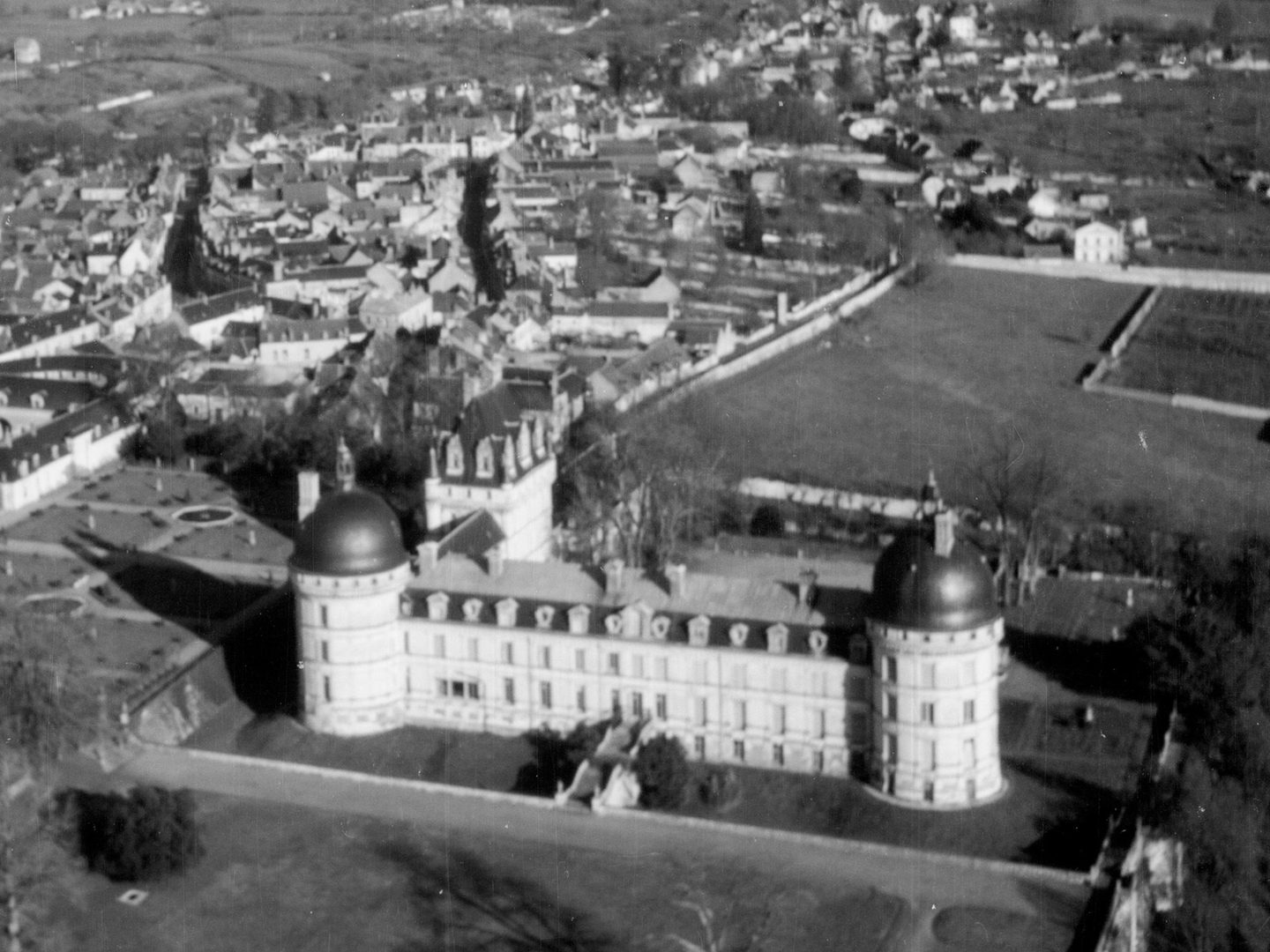 A historic black and white oblique aerial photo of a French chateau. It has white walls and 2 circular towers joined by a long wing. A second wing runs at rightangles from one of the towers to a square tower. It is surrounded by countryside with a town in the background.