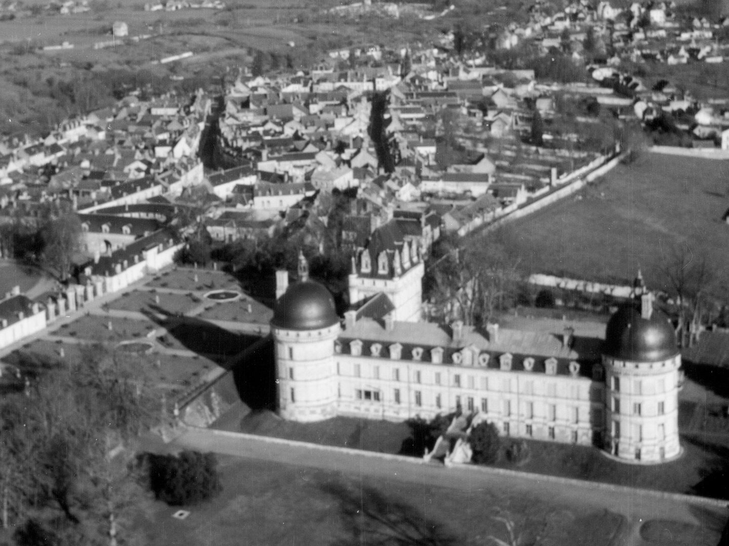 A historic black and white oblique aerial photo of a French chateau. It has white walls and 2 circular towers joined by a long wing. A second wing runs at rightangles from one of the towers to a square tower. It is surrounded by countryside with a town in the background.