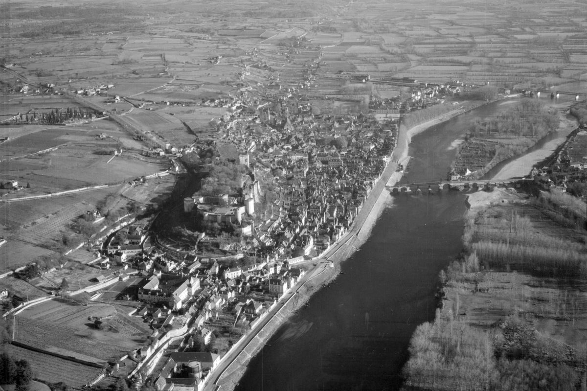 A black and white oblique aerial photo of a castle on a hill in a small town. A large river runs alongside the town and is crossed by a stone bridge. A landscape of fields and woods stretches into the distance.