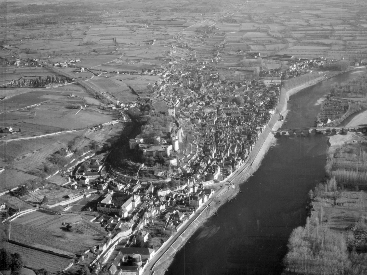 A black and white oblique aerial photo of a castle on a hill in a small town. A large river runs alongside the town and is crossed by a stone bridge. A landscape of fields and woods stretches into the distance.