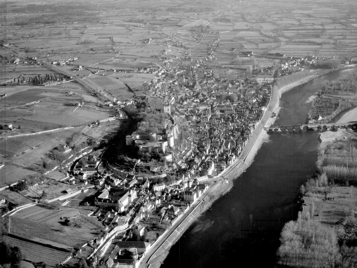 A black and white oblique aerial photo of a castle on a hill in a small town. A large river runs alongside the town and is crossed by a stone bridge. A landscape of fields and woods stretches into the distance.