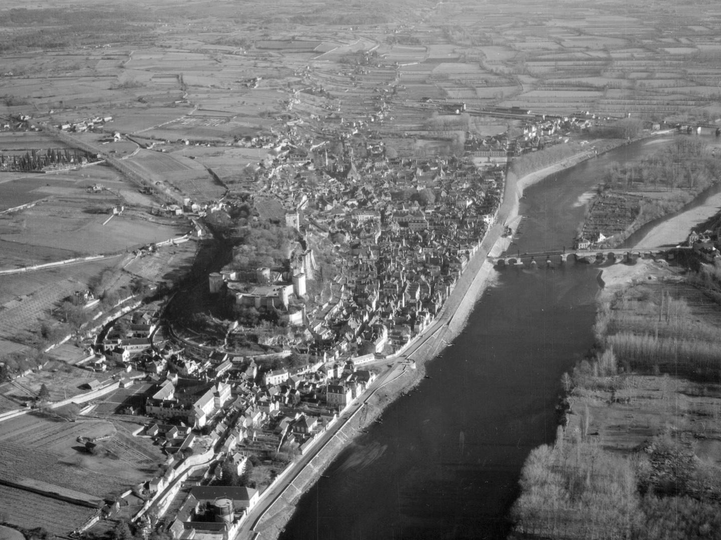 A black and white oblique aerial photo of a castle on a hill in a small town. A large river runs alongside the town and is crossed by a stone bridge. A landscape of fields and woods stretches into the distance.