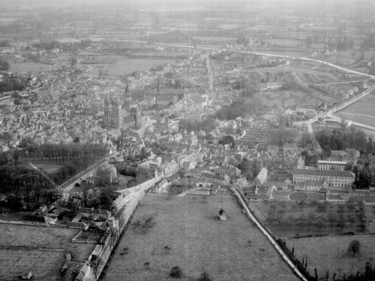 A black and white oblique aerial photo of a town fringed by fields and trees. A large cathedral dominates the centre of the town.