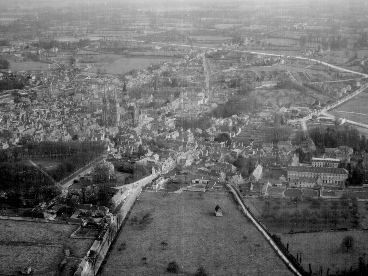 A black and white oblique aerial photo of a town fringed by fields and trees. A large cathedral dominates the centre of the town.