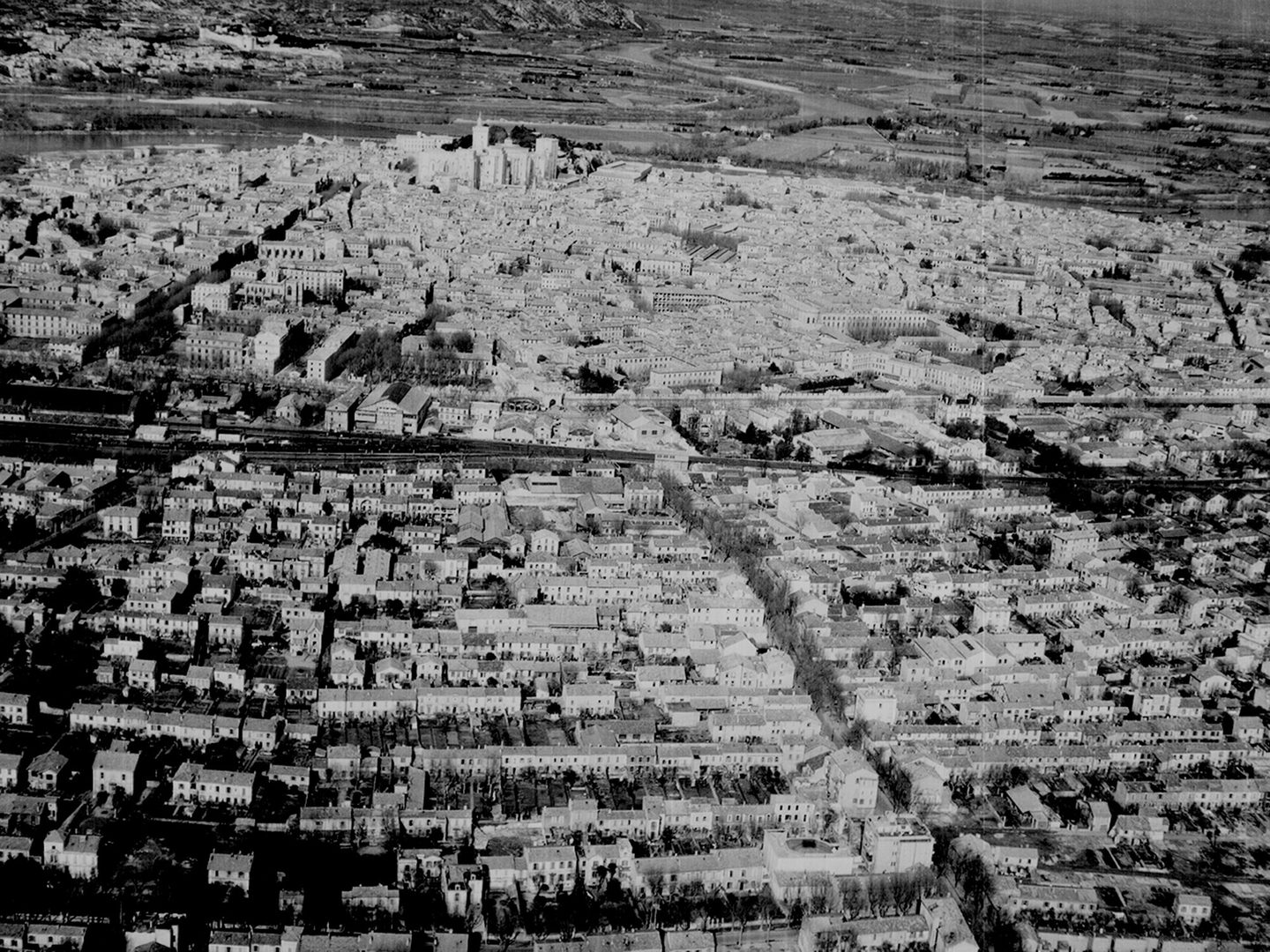 A historic black and white oblique aerial photo of a city. Houses rise up a slope towards a collection of grand buildings on the top of a hill. A watery landscape is visible beyond the grand buildings.