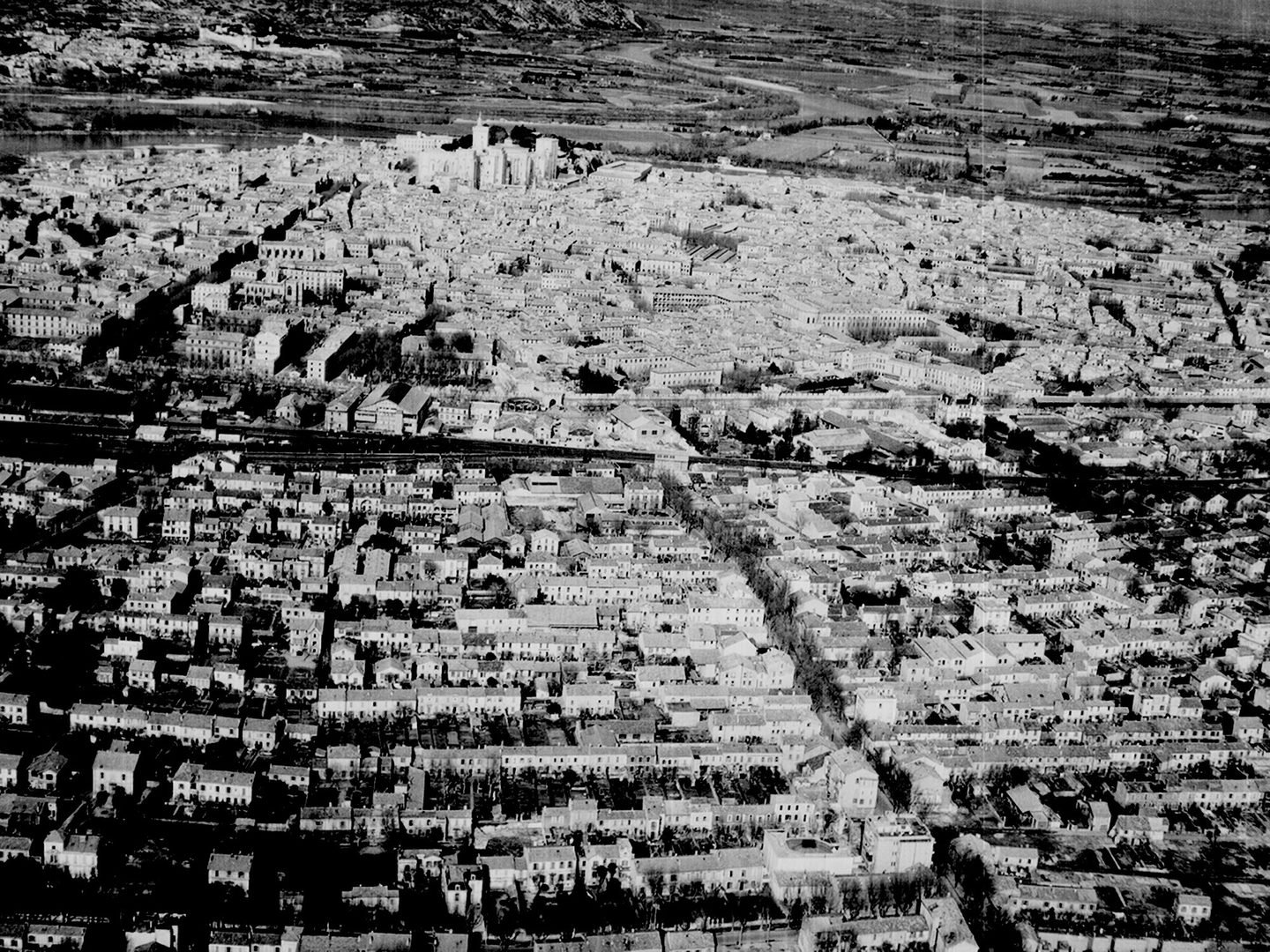 A historic black and white oblique aerial photo of a city. Houses rise up a slope towards a collection of grand buildings on the top of a hill. A watery landscape is visible beyond the grand buildings.
