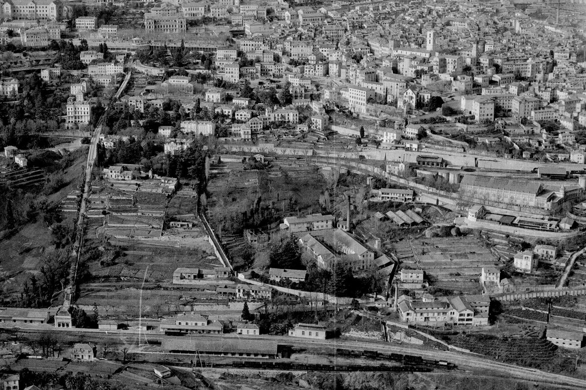 A historic black and white oblique aerial photo of a town climbing up the slope of a hill. The houses look Mediterranean. A railway runs through a station at the bottom of the hill, with lines of wagons visible on the tracks.
