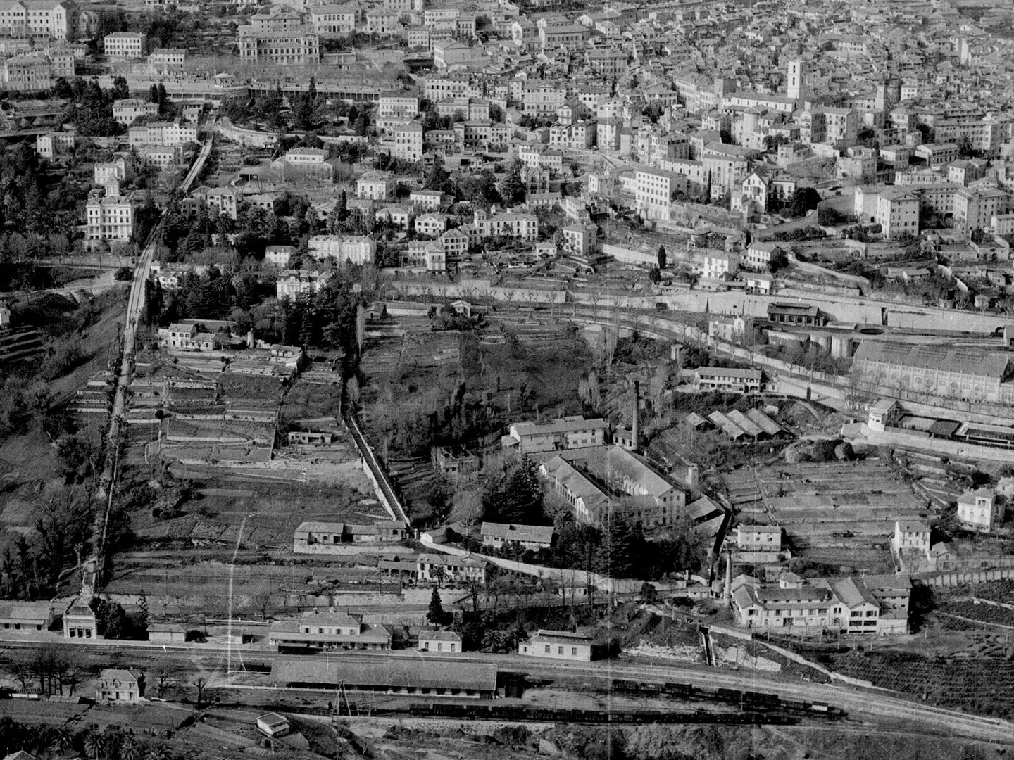 A historic black and white oblique aerial photo of a town climbing up the slope of a hill. The houses look Mediterranean. A railway runs through a station at the bottom of the hill, with lines of wagons visible on the tracks.