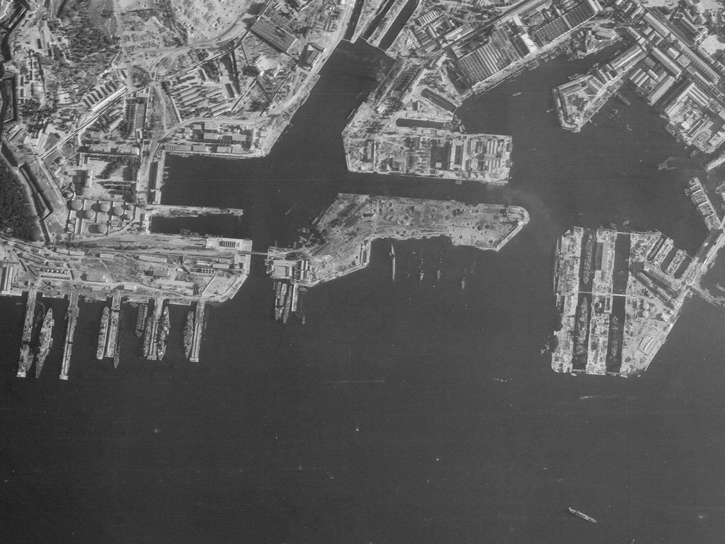 A historic black and white vertical aerial photo of a port. The docks run along the upper edge of the image with the sea along the lower edge. Numerous vessels are tied up at the docks, and some sunken vessels are also visible.