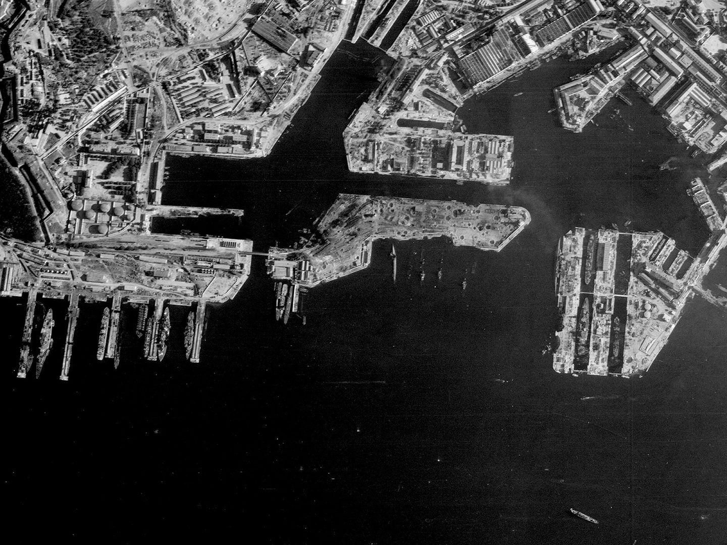 A historic black and white vertical aerial photo of a port. The docks run along the upper edge of the image with the sea along the lower edge. Numerous vessels are tied up at the docks, and some sunken vessels are also visible.