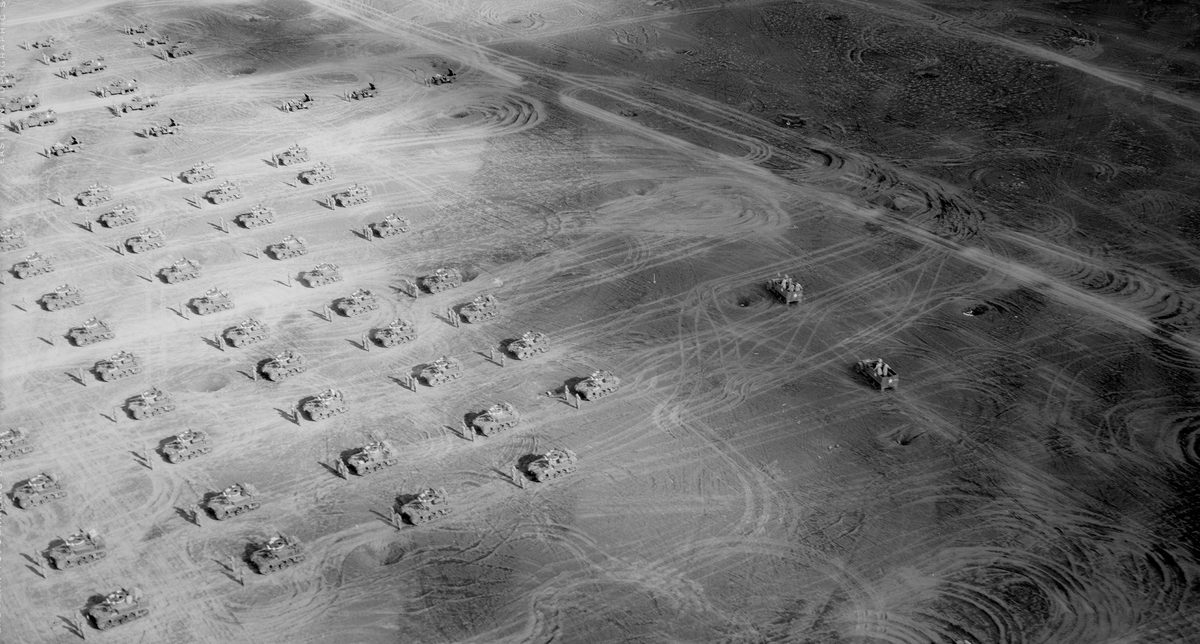 A historic black and white oblique aerial photo of a dusty landscape. The landscape is marked with numerous vehicle tracks. Arranged across it in a grid formation are tanks, each accompanied by a pair of soldiers.