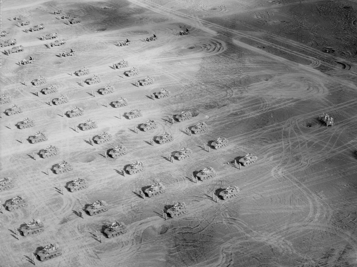 A historic black and white oblique aerial photo of a dusty landscape. The landscape is marked with numerous vehicle tracks. Arranged across it in a grid formation are tanks, each accompanied by a pair of soldiers.