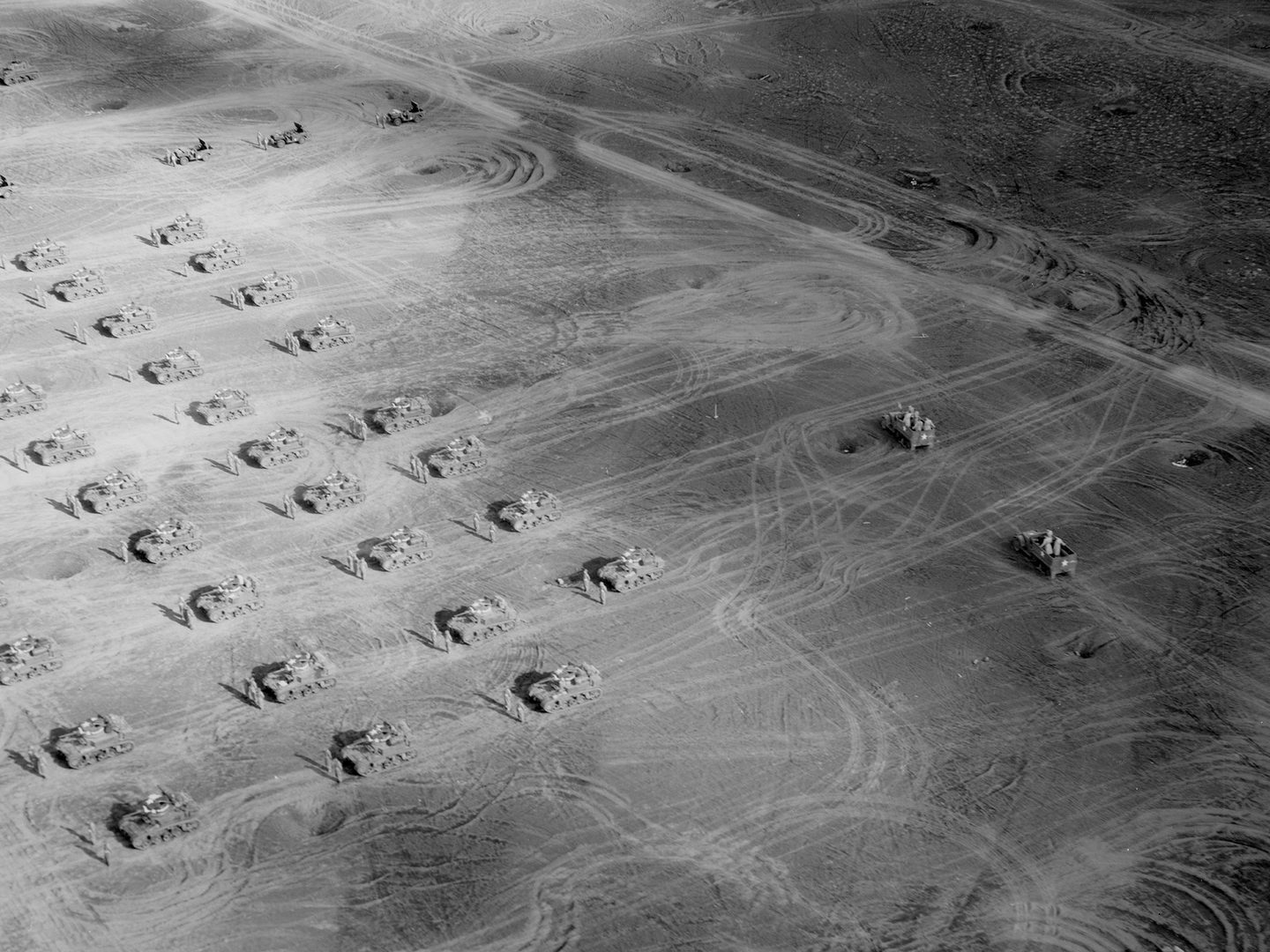 A historic black and white oblique aerial photo of a dusty landscape. The landscape is marked with numerous vehicle tracks. Arranged across it in a grid formation are tanks, each accompanied by a pair of soldiers.