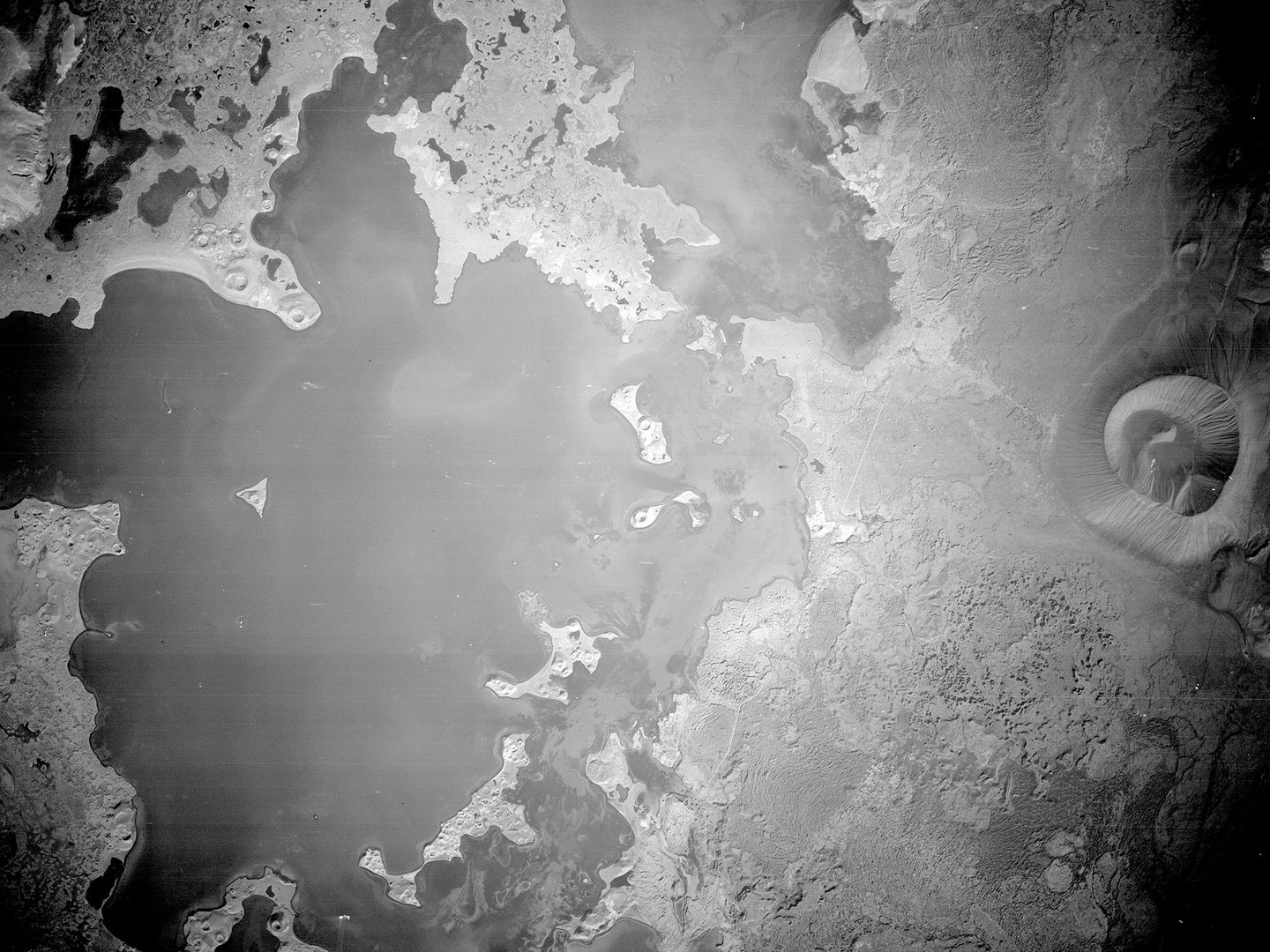 A black and white aerial photo of a large lake. It has irregular, serrated edges and is dotted with islands. To the right is a conical mountain with fluted sides and a large crater at the top.