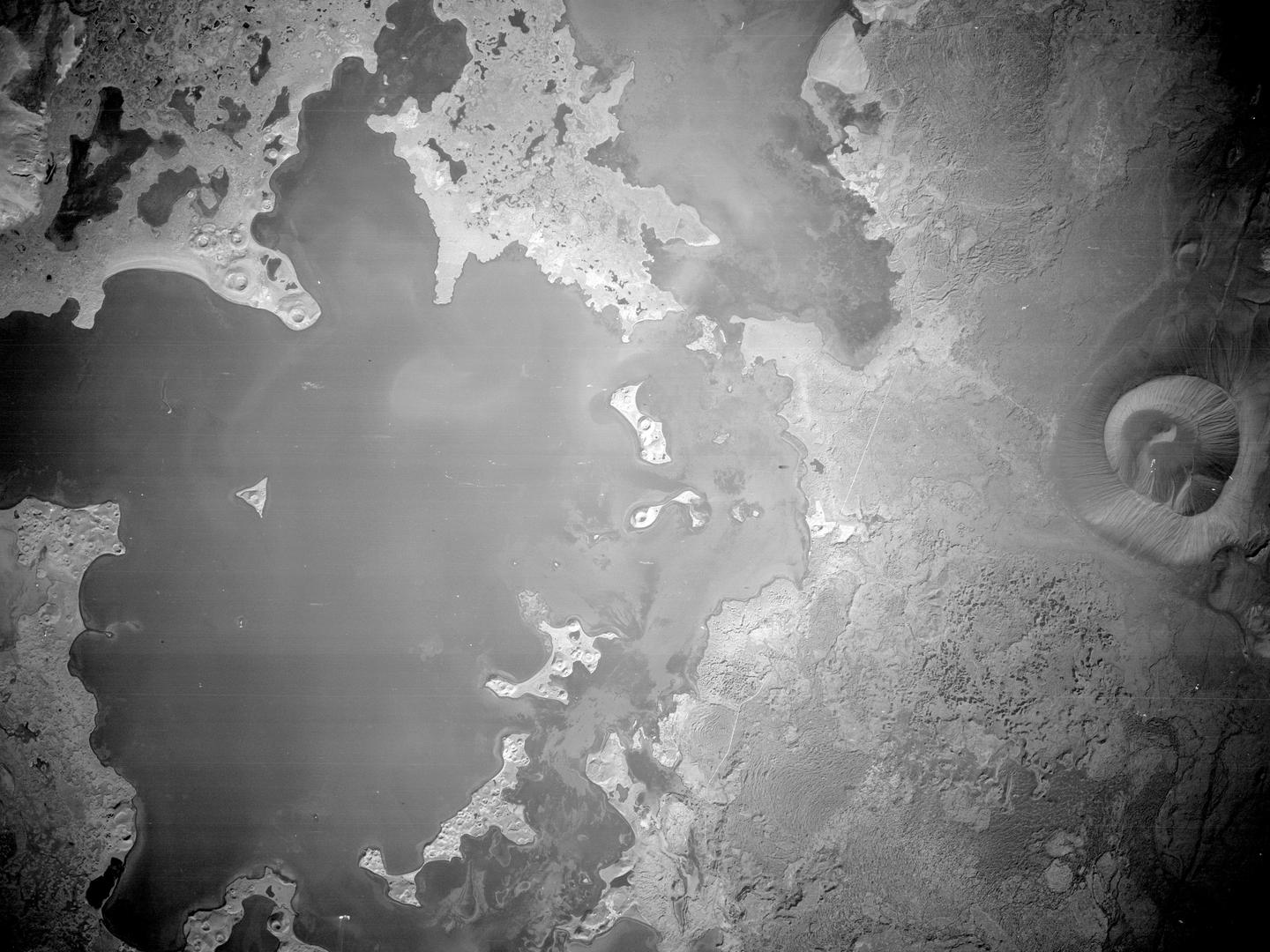 A black and white aerial photo of a large lake. It has irregular, serrated edges and is dotted with islands. To the right is a conical mountain with fluted sides and a large crater at the top.