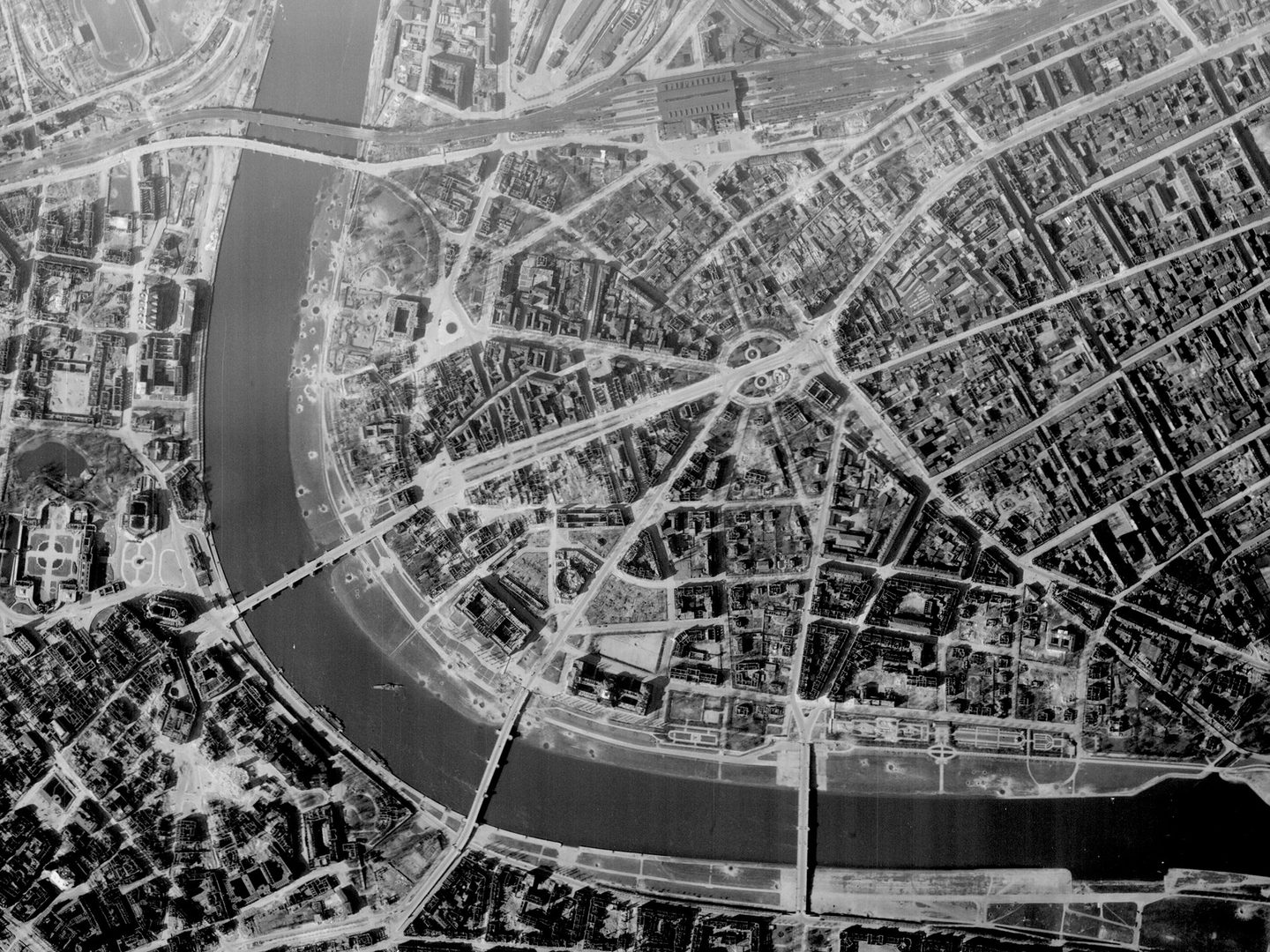 A black and white vertical aerial photo of Dresden. A wide river curves along the left and bottom edges, crossed by 4 bridges. Bomb damage is visible in roofless houses and craters.