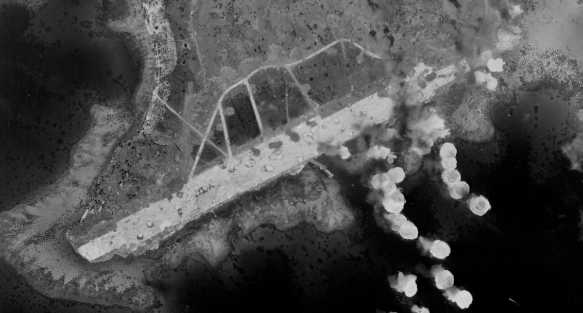 A historic black and white vertical aerial photo of an island headland, flanked by dark sea. A white runway extends along the headland. White flashes of explosions dot the righthand side of the image.