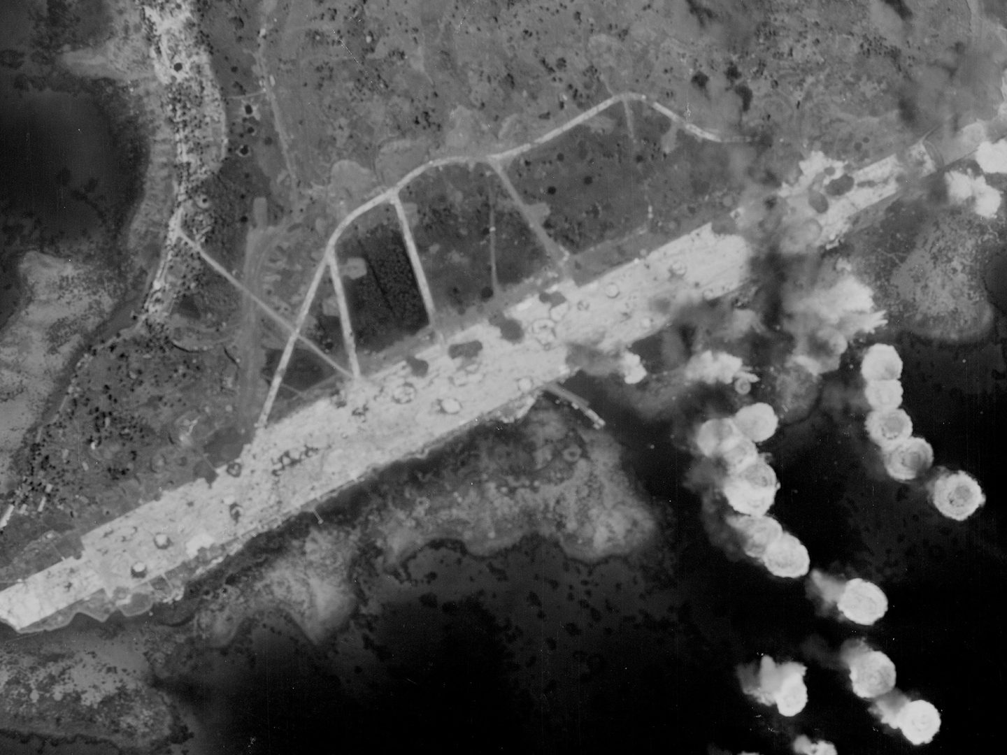 A historic black and white vertical aerial photo of an island headland, flanked by dark sea. A white runway extends along the headland. White flashes of explosions dot the righthand side of the image.