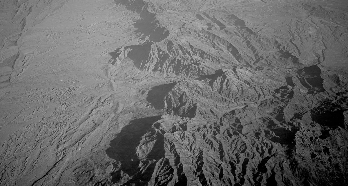 A historic black and white oblique aerial photo of a rugged, mountainous landscape. To the left, the mountains drop sharply to an arid plain.