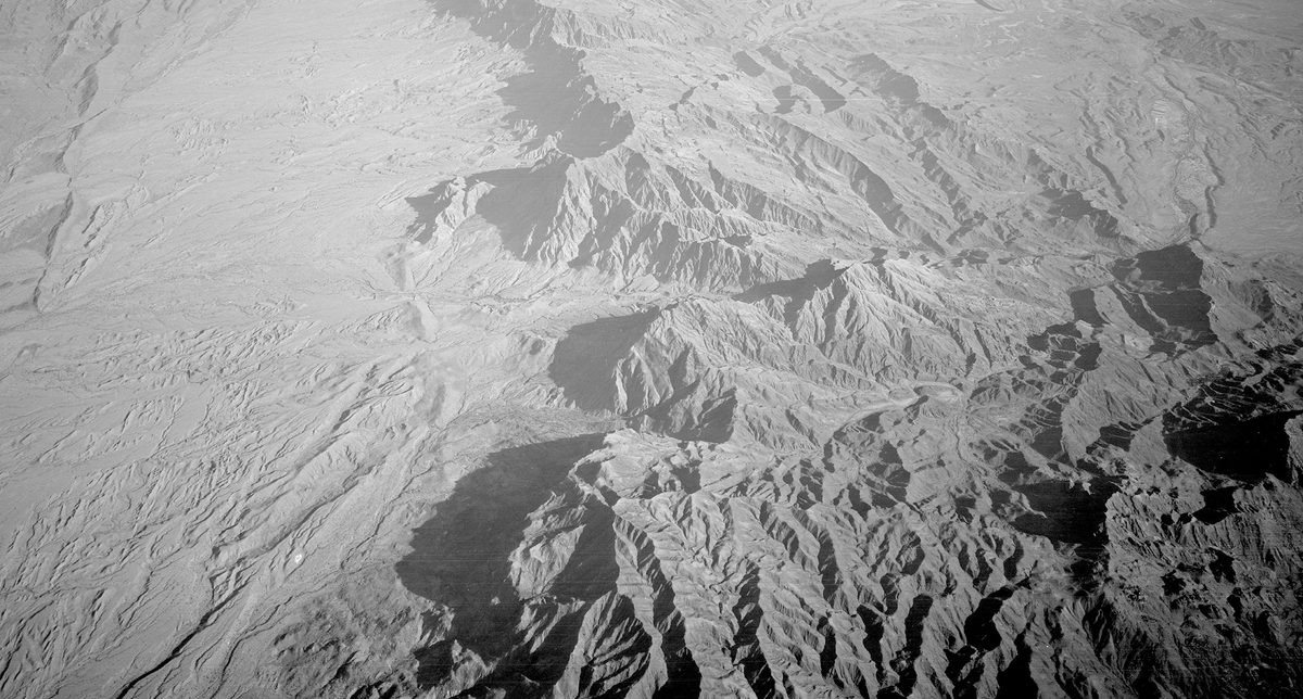 A historic black and white oblique aerial photo of a rugged, mountainous landscape. To the left, the mountains drop sharply to an arid plain.