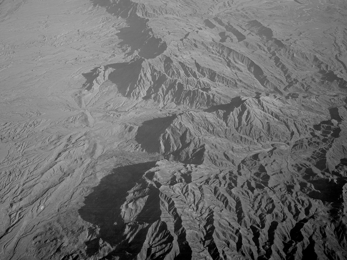A historic black and white oblique aerial photo of a rugged, mountainous landscape. To the left, the mountains drop sharply to an arid plain.