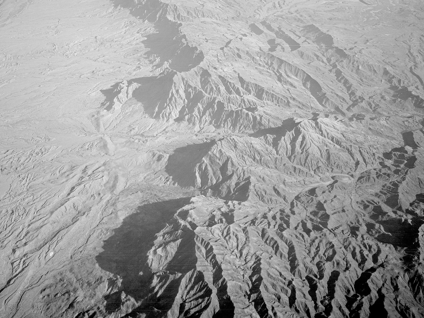 A historic black and white oblique aerial photo of a rugged, mountainous landscape. To the left, the mountains drop sharply to an arid plain.