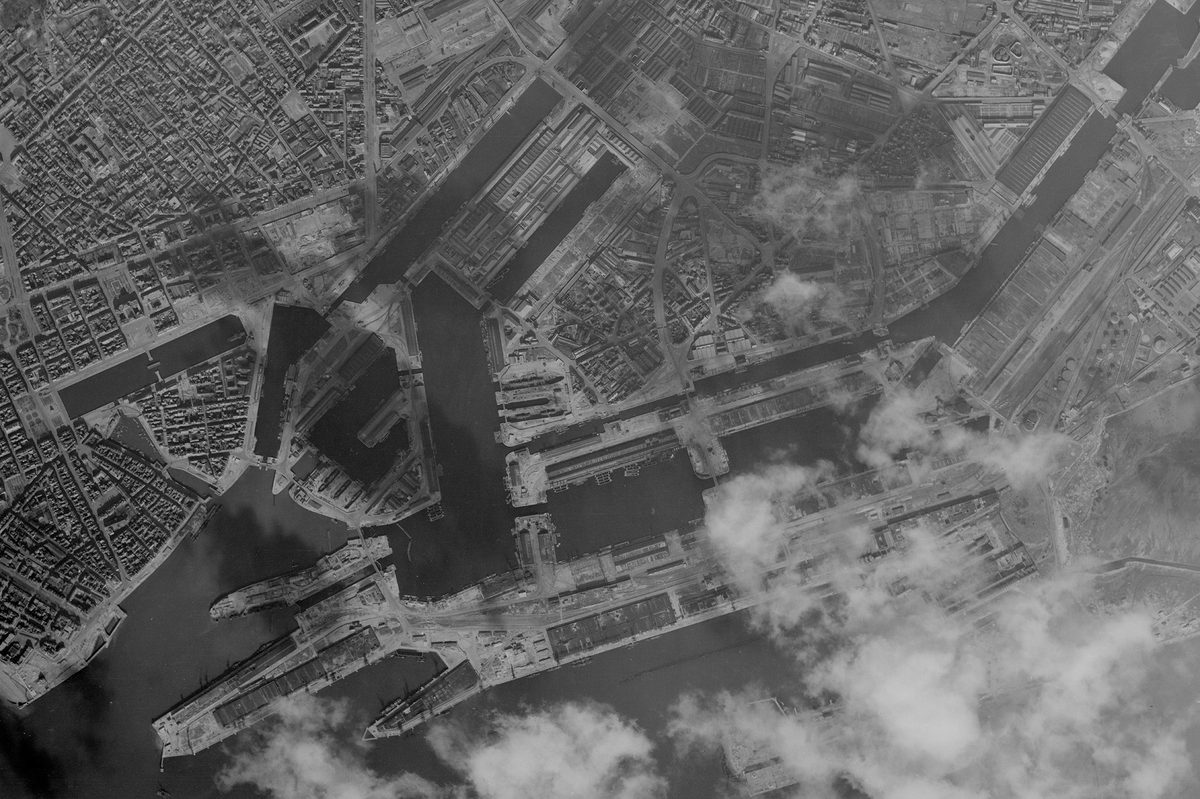 A historic black and white vertical aerial photo of a port. Numerous docks stretch into the surrounding city. Clouds obscure part of the image at the bottom right.