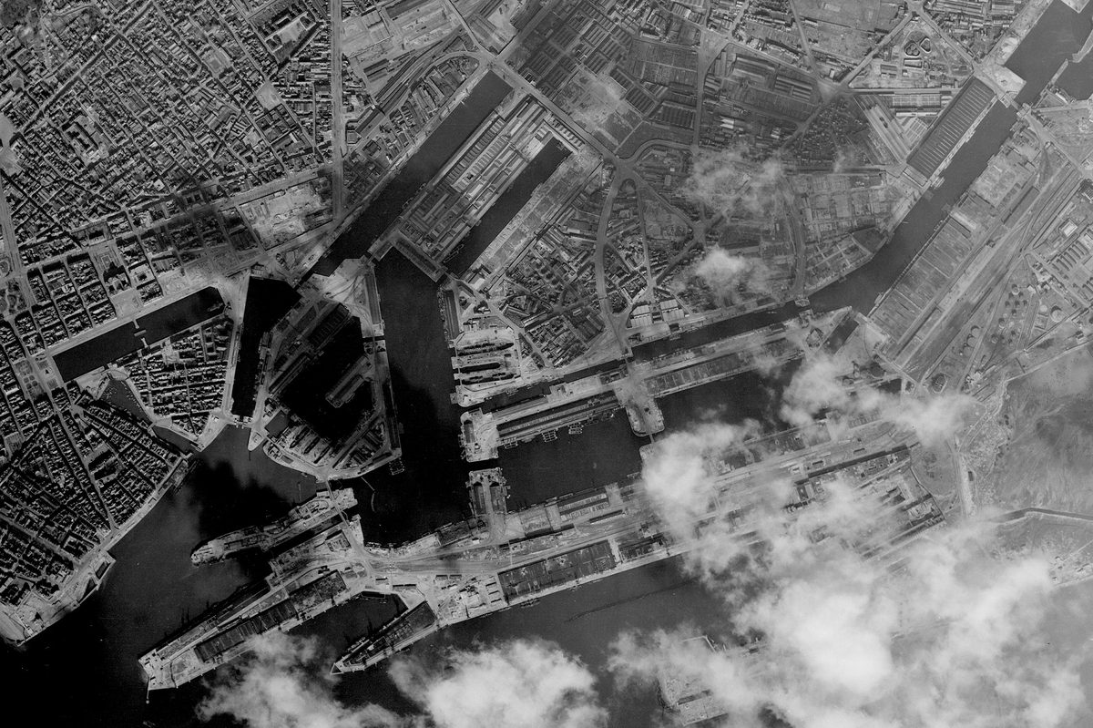 A historic black and white vertical aerial photo of a port. Numerous docks stretch into the surrounding city. Clouds obscure part of the image at the bottom right.