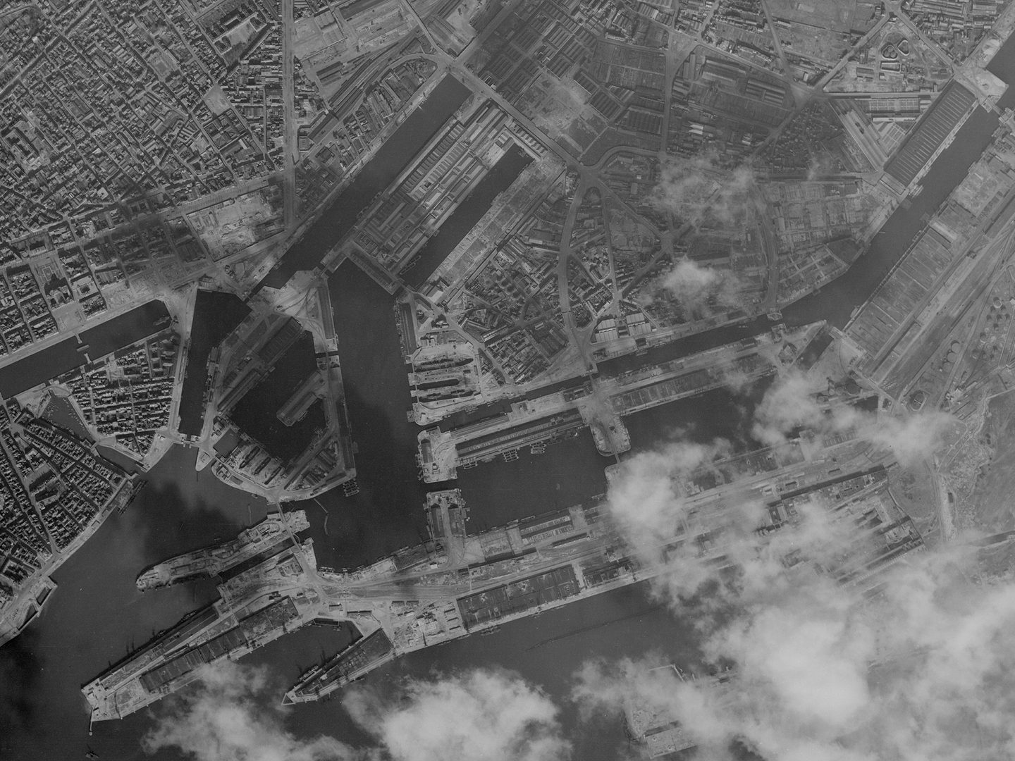 A historic black and white vertical aerial photo of a port. Numerous docks stretch into the surrounding city. Clouds obscure part of the image at the bottom right.