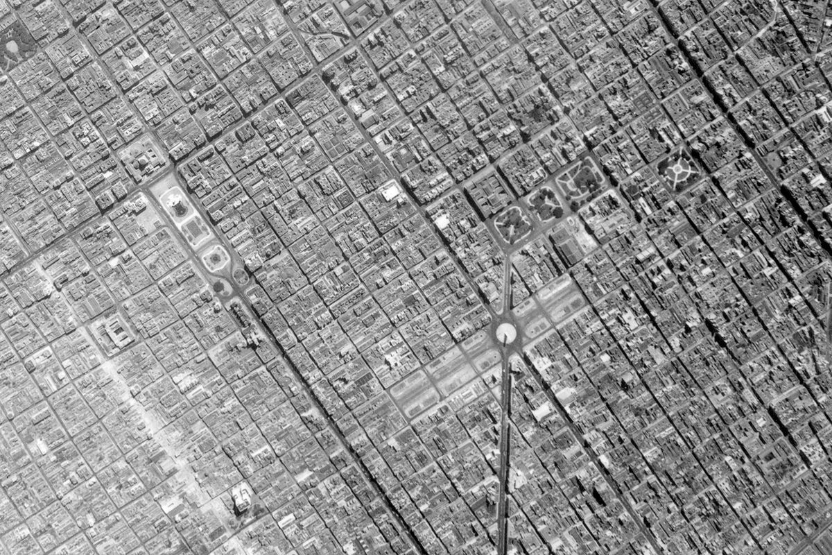 A historic black and white vertical aerial photo of a large city broken up into a regular grid. Just below right of the centre is a monument in the middle of a wide boulevard.