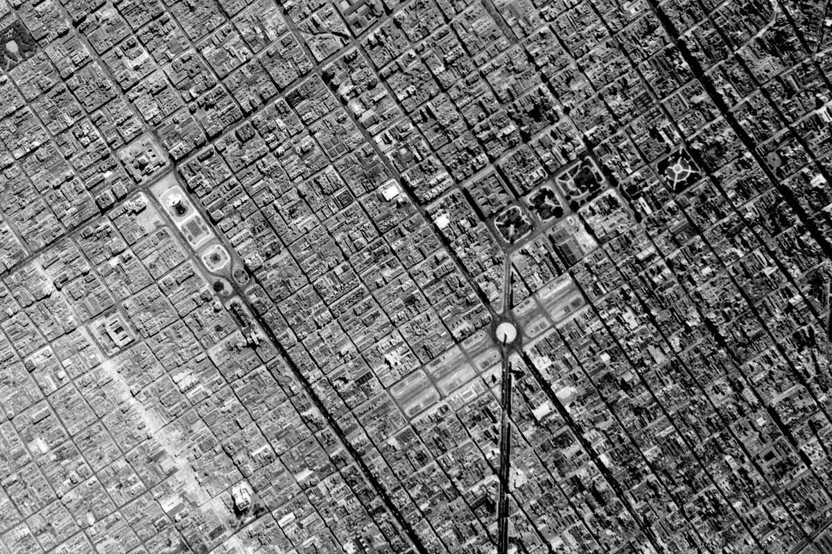A historic black and white vertical aerial photo of a large city broken up into a regular grid. Just below right of the centre is a monument in the middle of a wide boulevard.