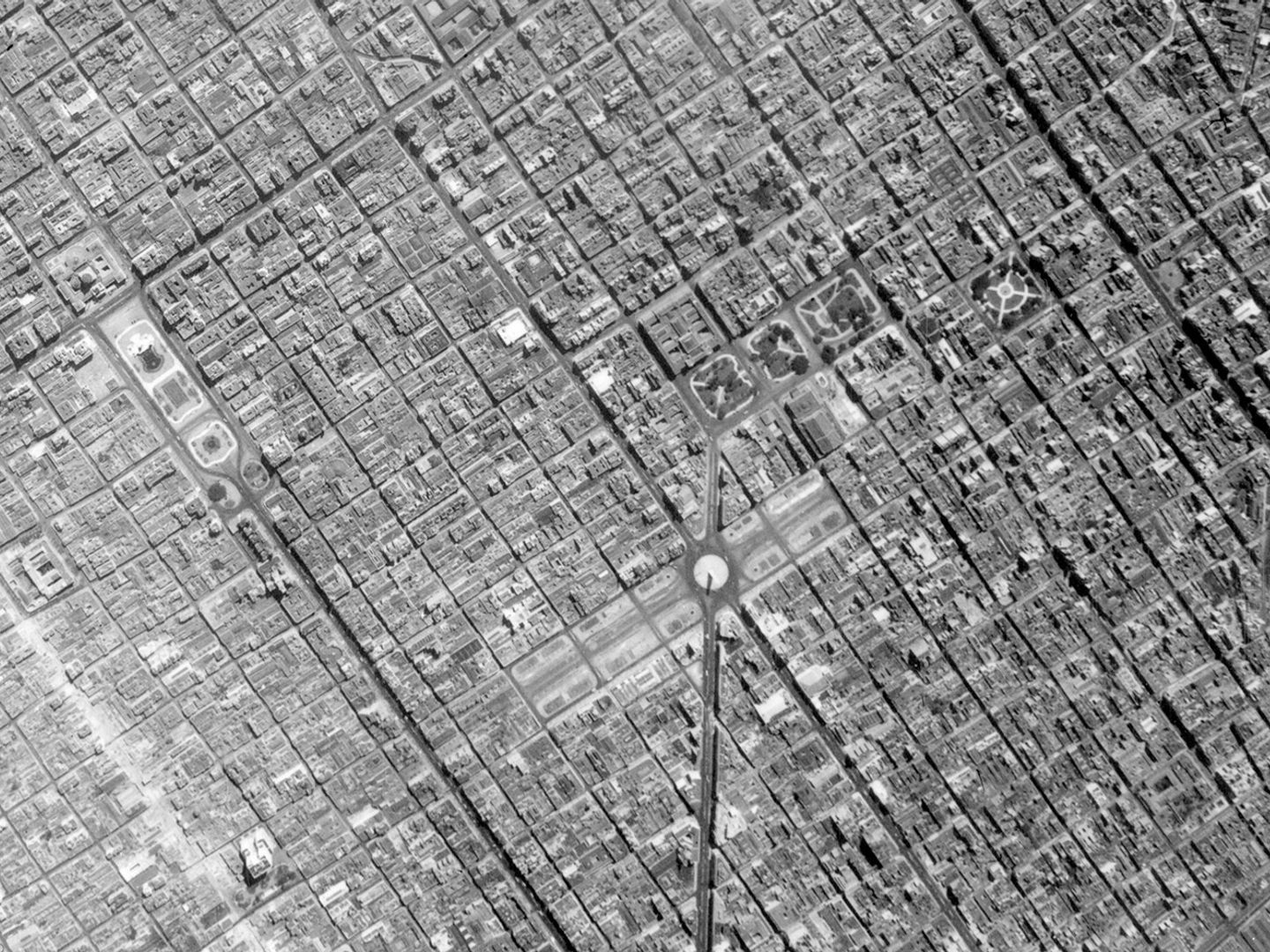 A historic black and white vertical aerial photo of a large city broken up into a regular grid. Just below right of the centre is a monument in the middle of a wide boulevard.