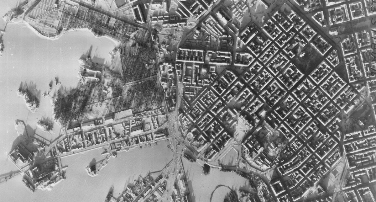 A historic black and white vertical aerial photo of Helsinki in winter. The regular grid of streets stands out against the snow. To the left is a ragged coastline fringed by frozen sea.