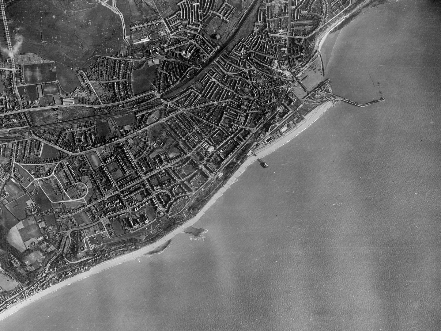 A historic black and white vertical aerial photo of a town on a coast. The town stretches along the coastline along the top and left-hand edges of the image. A pier projects into the sea towards the top right. The sea fills the bottom right half of the image.