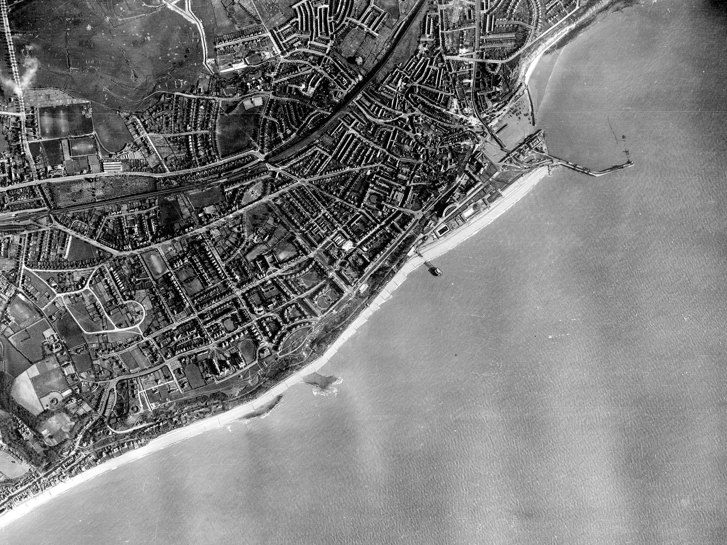A historic black and white vertical aerial photo of a town on a coast. The town stretches along the coastline along the top and left-hand edges of the image. A pier projects into the sea towards the top right. The sea fills the bottom right half of the image.