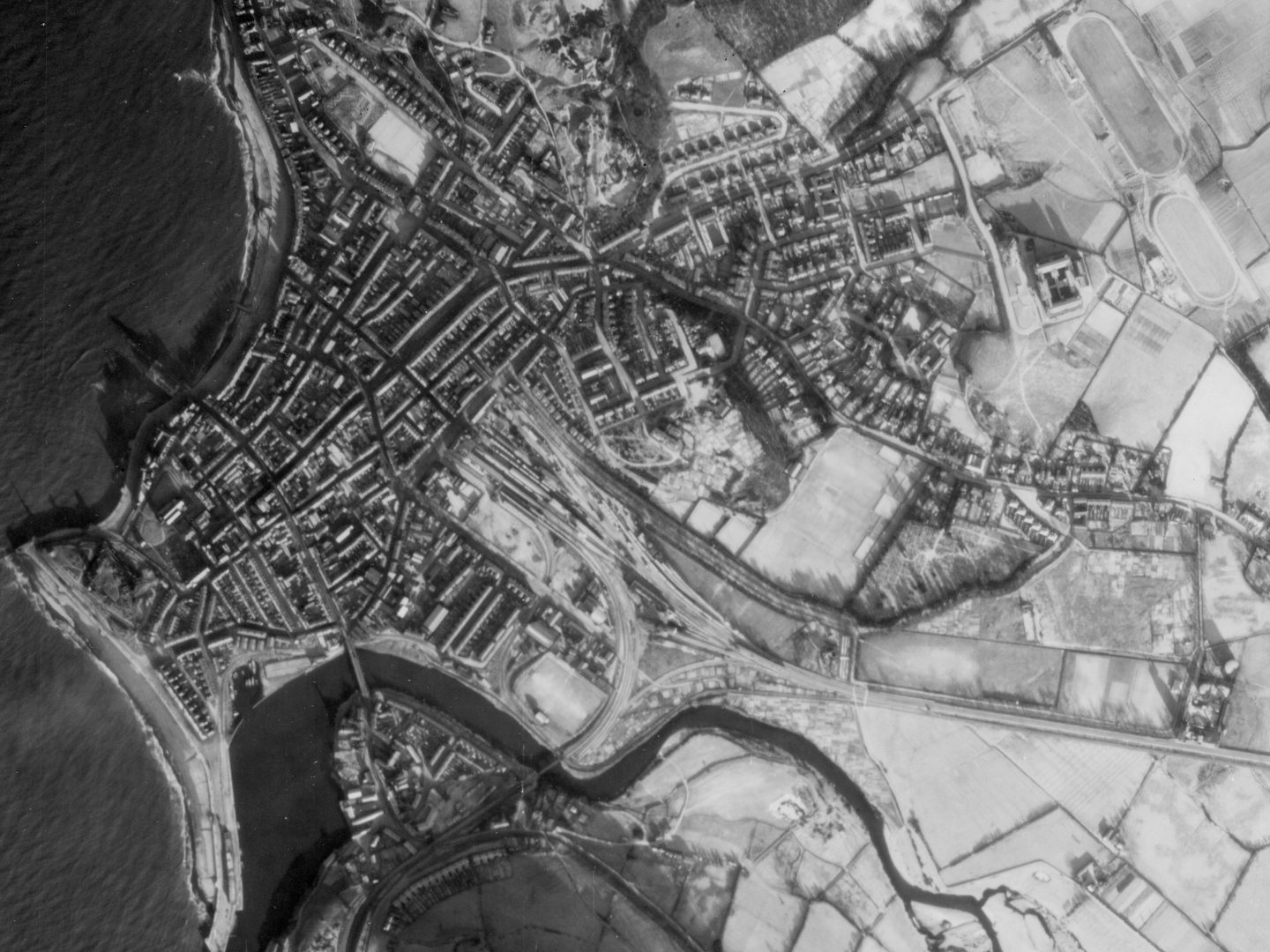 A historic black and white vertical aerial photo of the town of Aberystwyth, with the sea to the left and the town to the right. A river runs along the bottom of the image. The extensive fields to the right of the image are covered in snow.