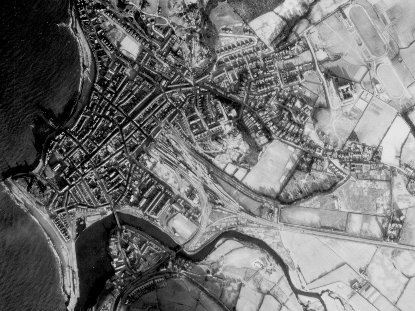 A historic black and white vertical aerial photo of the town of Aberystwyth, with the sea to the left and the town to the right. A river runs along the bottom of the image. The extensive fields to the right of the image are covered in snow.