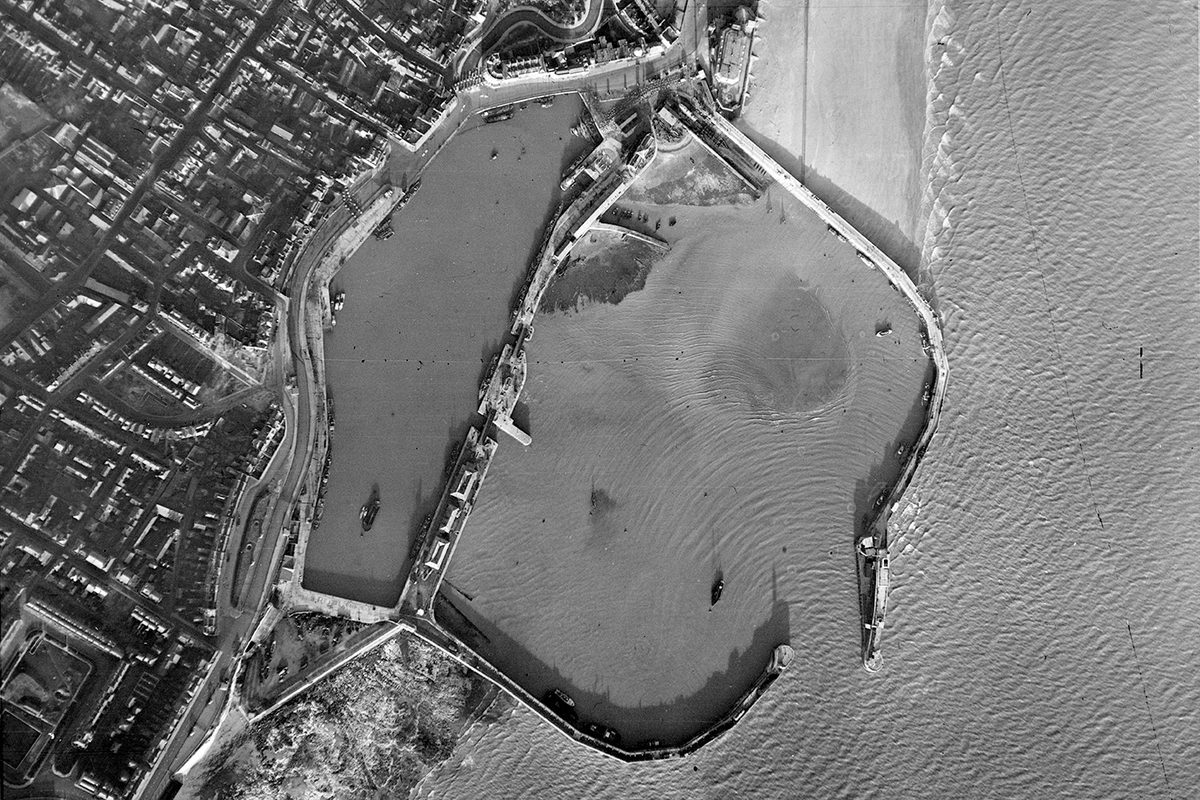 A historic black and white vertical aerial photo of a small harbour. The walls of the harbour reach to the right from the land into the sea, almost meeting and leaving only a narrow entrance for boats. A town is visible on the shore to the left.