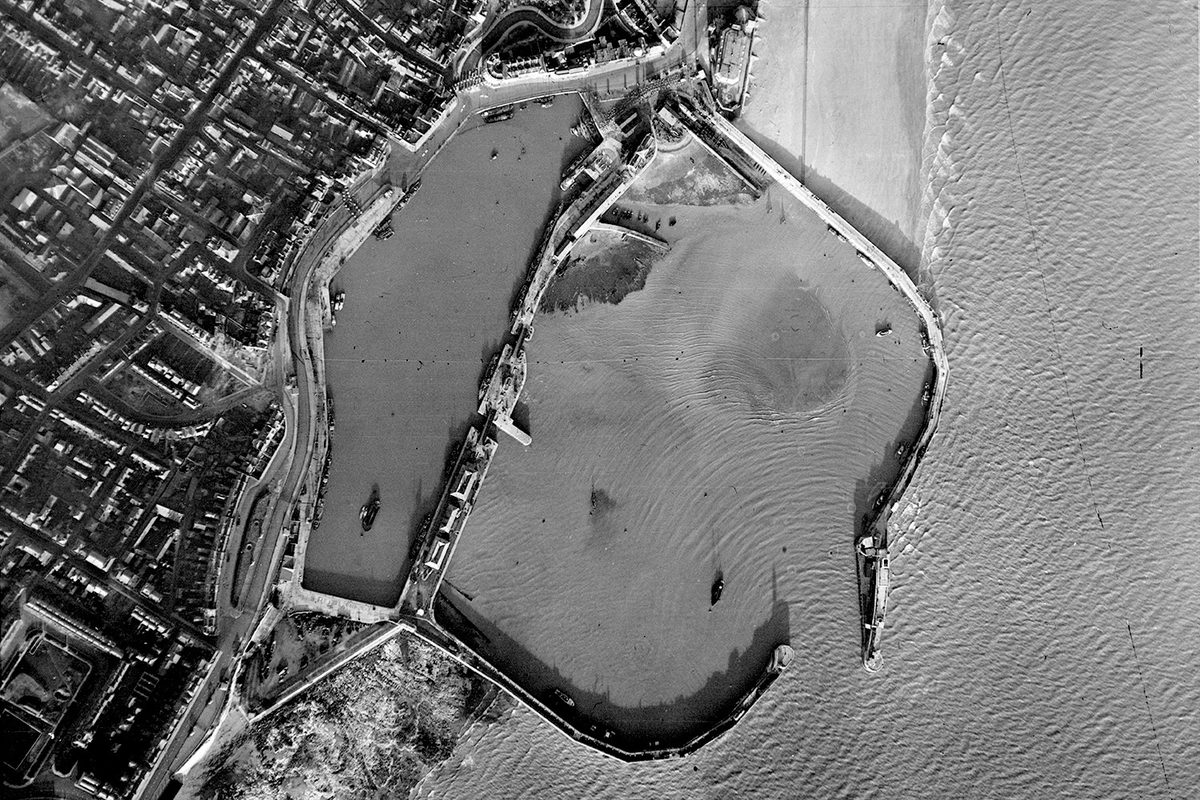 A historic black and white vertical aerial photo of a small harbour. The walls of the harbour reach to the right from the land into the sea, almost meeting and leaving only a narrow entrance for boats. A town is visible on the shore to the left.