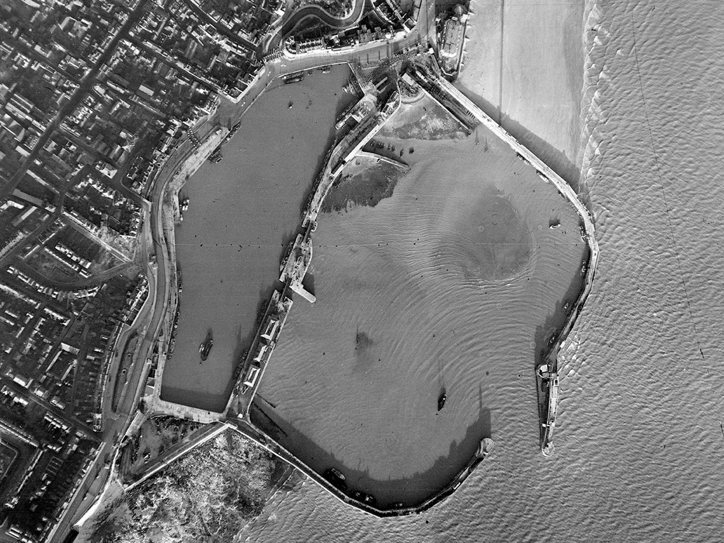 A historic black and white vertical aerial photo of a small harbour. The walls of the harbour reach to the right from the land into the sea, almost meeting and leaving only a narrow entrance for boats. A town is visible on the shore to the left.